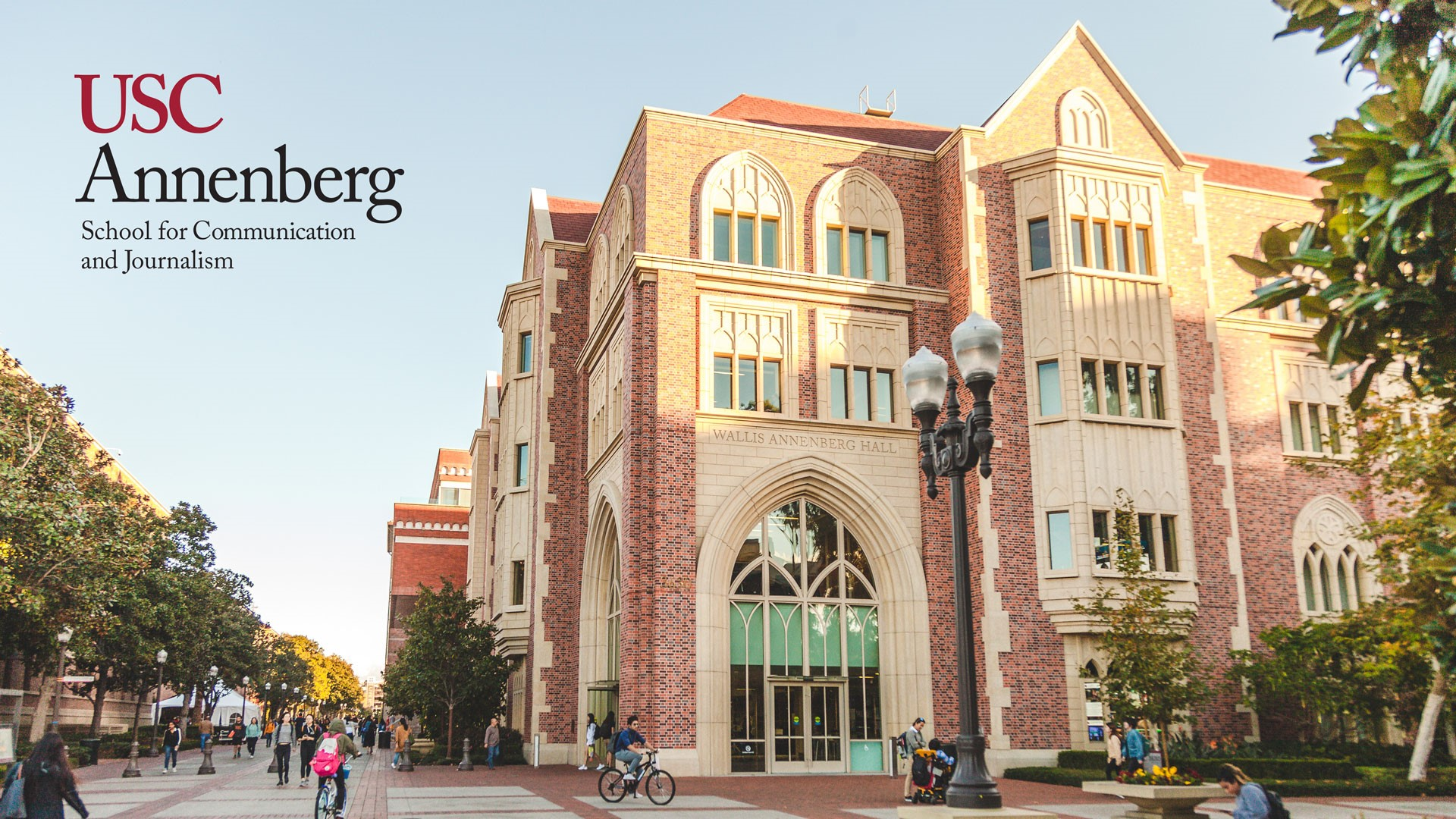 A photo of an imposing brick and masonry building with gothic and Spanish influences and a wide campus lane with cement and paver stones lined with trees. The USC Annenberg School for Communication and Journalism logo in red and black sits at the top left.