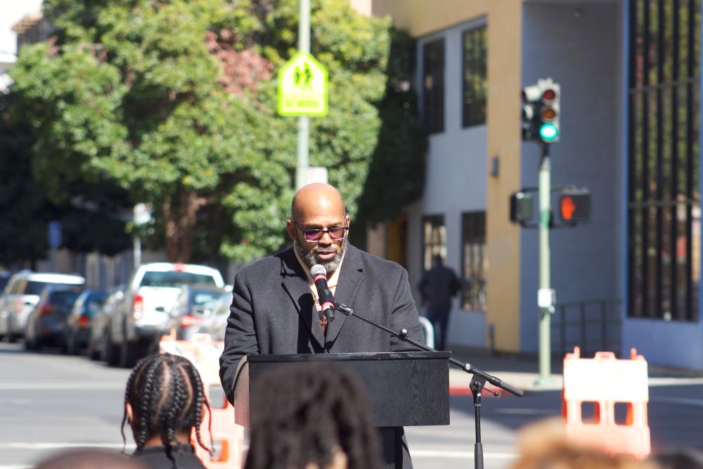 A photo of Martin G. Reynolds, a Black man, bald, wearing tinted glasses and a wool coat, stands at a podium with a mic stand on a street corner and addresses a small group of people who become blurry in the foreground.
