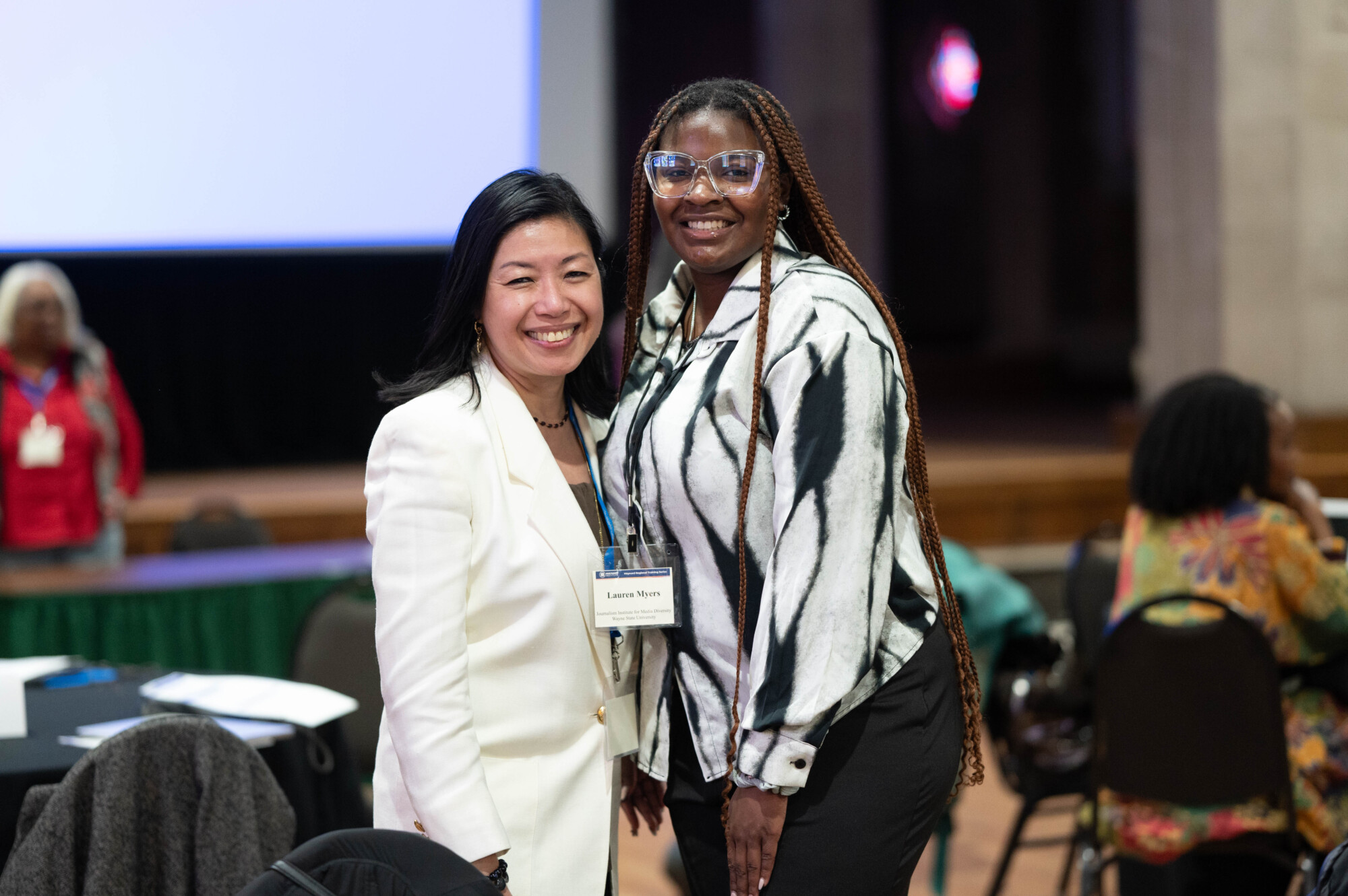 Photo of Odette Alcazaren-Keeley, left, an Asian woman with shoulder-length black hair and a white suit, posing and smiling with Lauren Myers, a young Black woman with a black and white shirt, lanyard and name tag, large clear eyeglasses, and long brown braids.