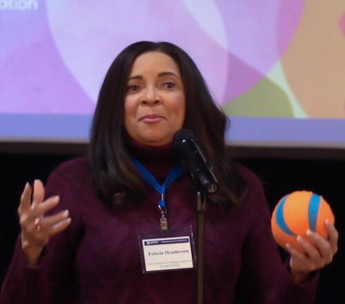 A photo of Felecia D. Henderson, a Black woman with a medium complexion and long dark hair. She wears a dark purple cable-knit sweater and a blue logo affixed with a name tag. She holds what appears to be a small orange and blue basketball. Behind her a projection screen shows a colorful collage.