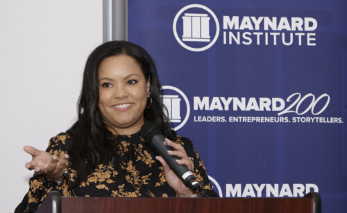 A young black woman with shoulder-length wavy hair wears a black blouse with a golden floral motif. She smiles and stands at a podium, holding a microphone. Behind her is a dark blue banner with the Maynard Institute logo in white.