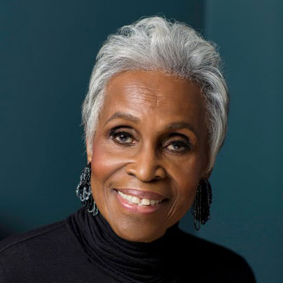 A photo of Dorothy Butler-Gilliam, an older Black woman with white hair, wearing a black turtleneck and dangling beaded earrings. Behind her is a dark teal background.