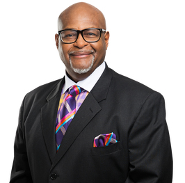 A photo of John X. Miller, a Black man with a graying goatee. He is bald and wears dark glasses and a black suit with a colorful shirt and matching tie and pocket square.