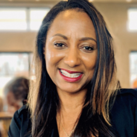 A photo of Susan Leath, a Black woman wearing a black blouse, reddish-pink lipstick, and with long hair in shades of dark to honey brown.