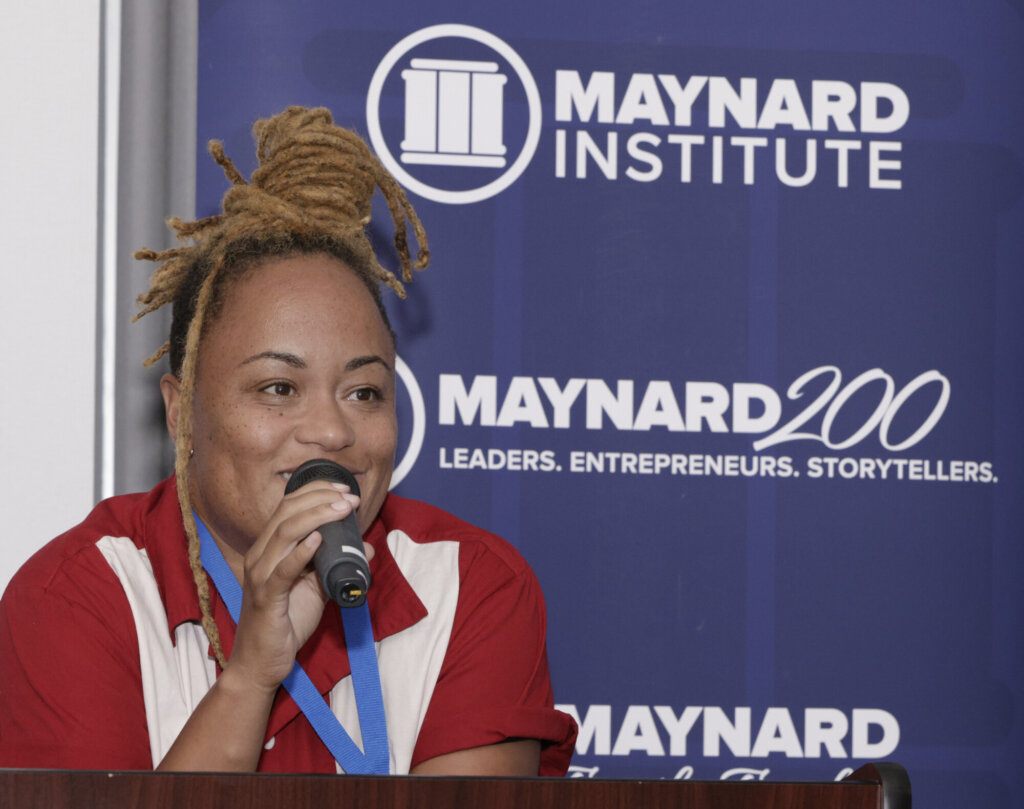 A photo of a young Black person standing in front of a Maynard Institute branded banner holding a microphone, light colored locks piled into a coiled bun atop their head.