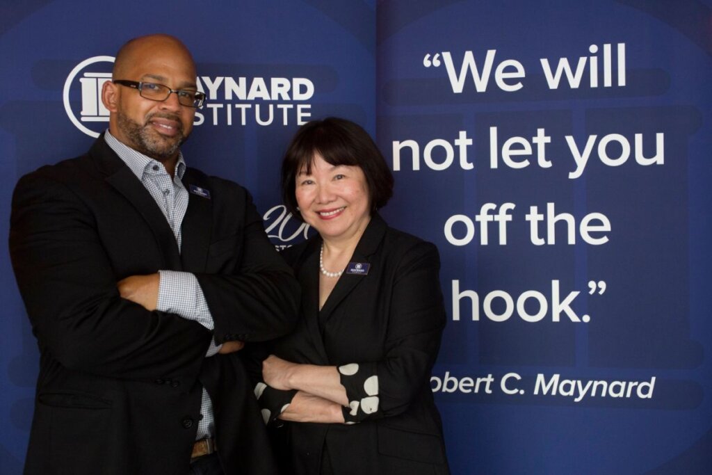 A photo of Martin G. Reynolds, a Black man, bald and wearing glasses and professional attire, standing before a dark blue banner with Maynard Institute branding. Next to him, Evelyn Hsu, an older Asian woman with a bob haircut and bangs wearing a black suit pearls. Beside her, part of the banner reads 