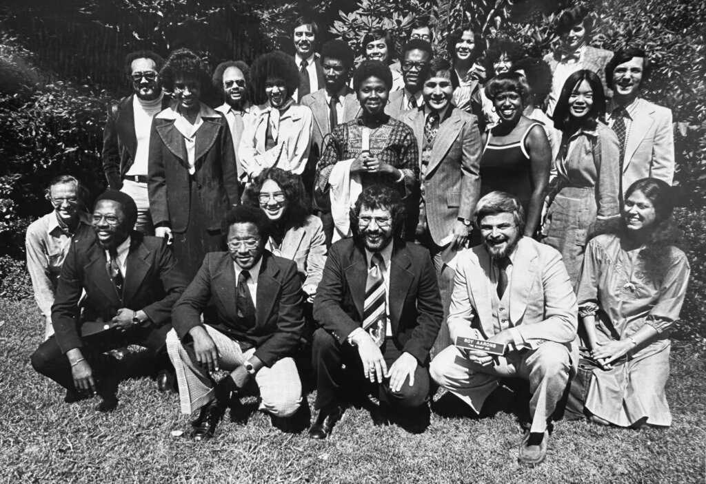 Over a dark blue background, a black and white photo of a group of people of various ages and races standing outdoors and posing for the camera in a group. The front row kneels. From their clothes, hair and glasses this looks to be the 1970s.
