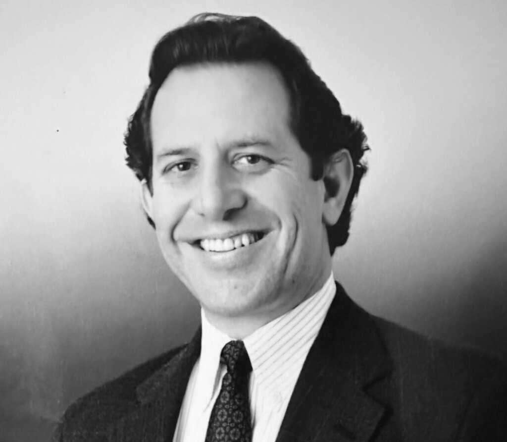 a black and white photo of Steve Montiel, a smiling man with dark wavy hair combed back and a dark suit and patterned tie.
