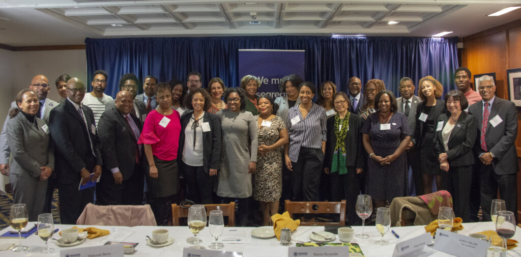 A group of more than two dozen people of different races and ages gathers for a group photo. Behind them, a deep blue curtain and a dark blue standing banner with Maynard Institute branding. In front of them in the foreground, a banquet table where it appears they have just been eating dinner.