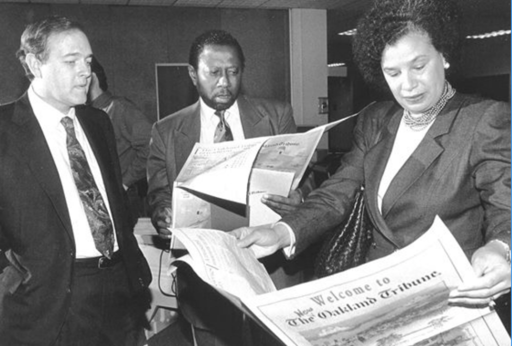 Over a dark-blue background: in the foreground, Nancy Hicks Maynard, a Black woman with dark curly hair graying at the temples, wears professional attire and holds a newspaper, the front page of which reads 