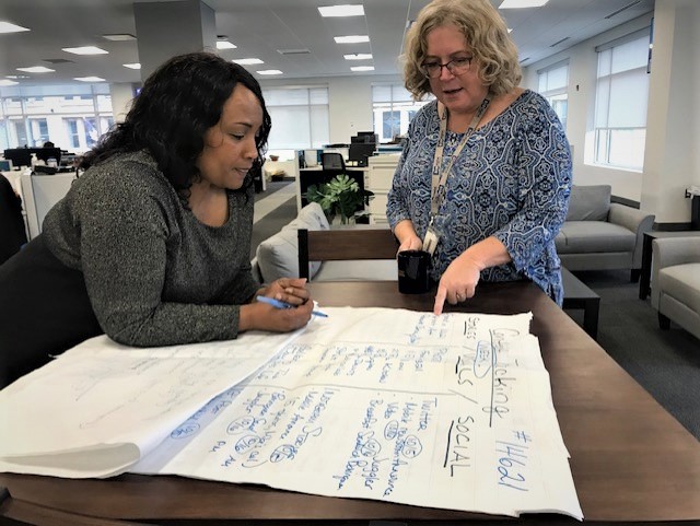 A Black woman and white woman lean over a large table and work together on a large poster in a corporate office setting. The Black woman at left holds a pen, the white woman at right holds a coffee mug and points at a section of the poster.
