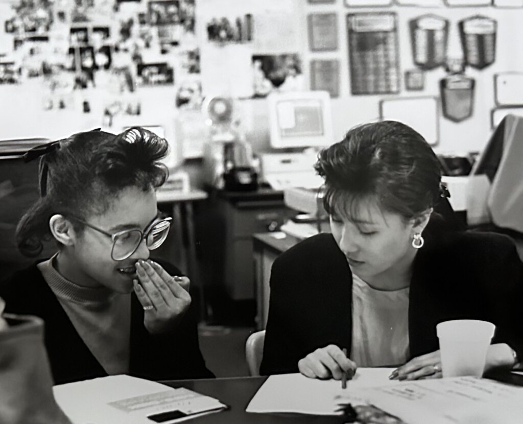 A black and white photo of two students, a young Black woman at left with large glasses and styled bangs, and a young Asian woman to the right wearing a black suit jacket and gold earrings. They collaborate on a document. Behind them old photos and trophy plaques line one wall and other computer desks are in view.