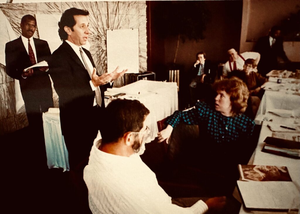 Steve Montiel, a man with wavy dark hair, addresses people seated at banquet tables. Their clothing and the setting denote the 1990s.