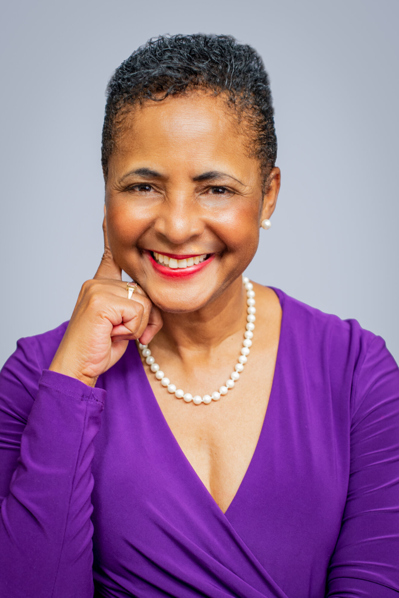 A headshot photo of Dorothy Bland, a Black woman with short hair and a bright smile, leaning her chin on her hand. She wears a bright purple blouse and pearl necklace.