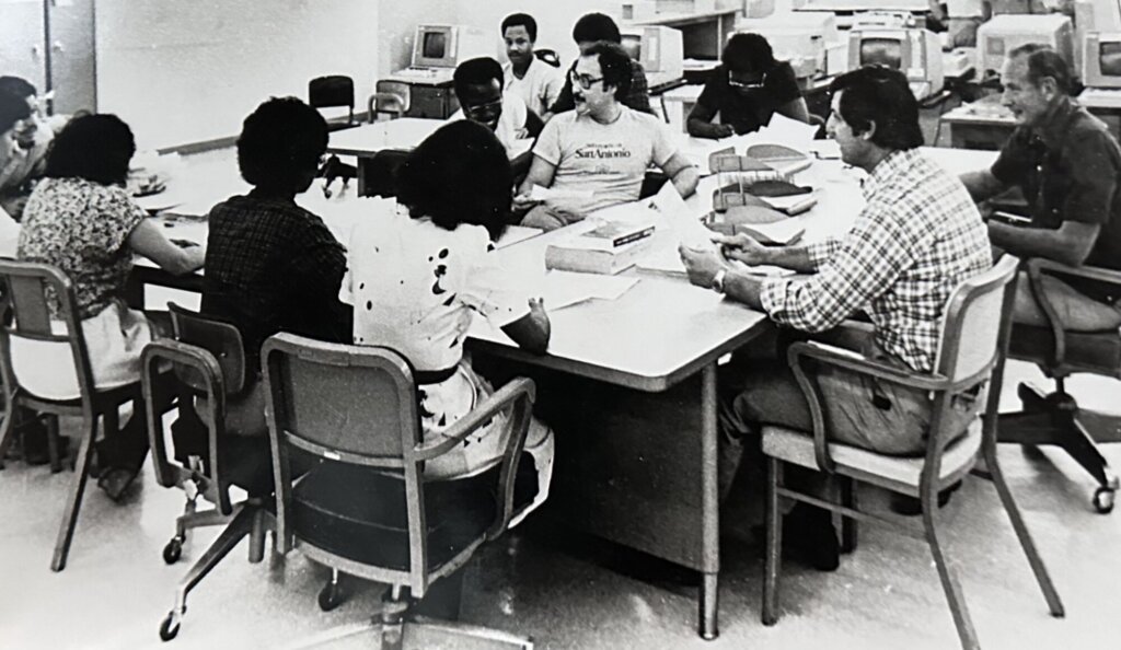 A black and white photo shows a group of men and women of different races in an office, sitting in chairs around tables that form a horseshoe. From their glasses, clothes, hairstyles, and the furniture and computers in the background, this photo appears to have been taken in the 1970s or early 1980s.