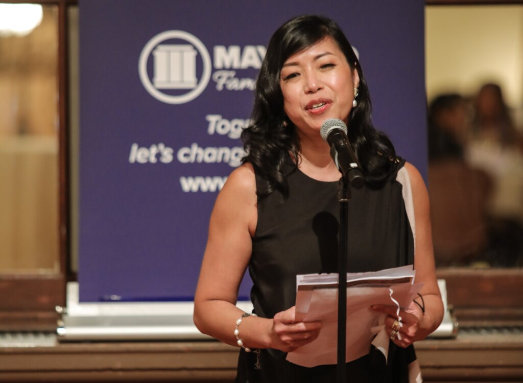 A photo of Odette Alcazaren-Keeley in evening/professional dress, wearing a black and white sleeveless dress and pearl bracelet and earrings. She is an Asian woman with dark shoulder-length wavy hair. Behind her is a dark blue standing banner with Maynard Institute branding. She speaks into a microphone and holds a stack of papers as though presenting a speech.