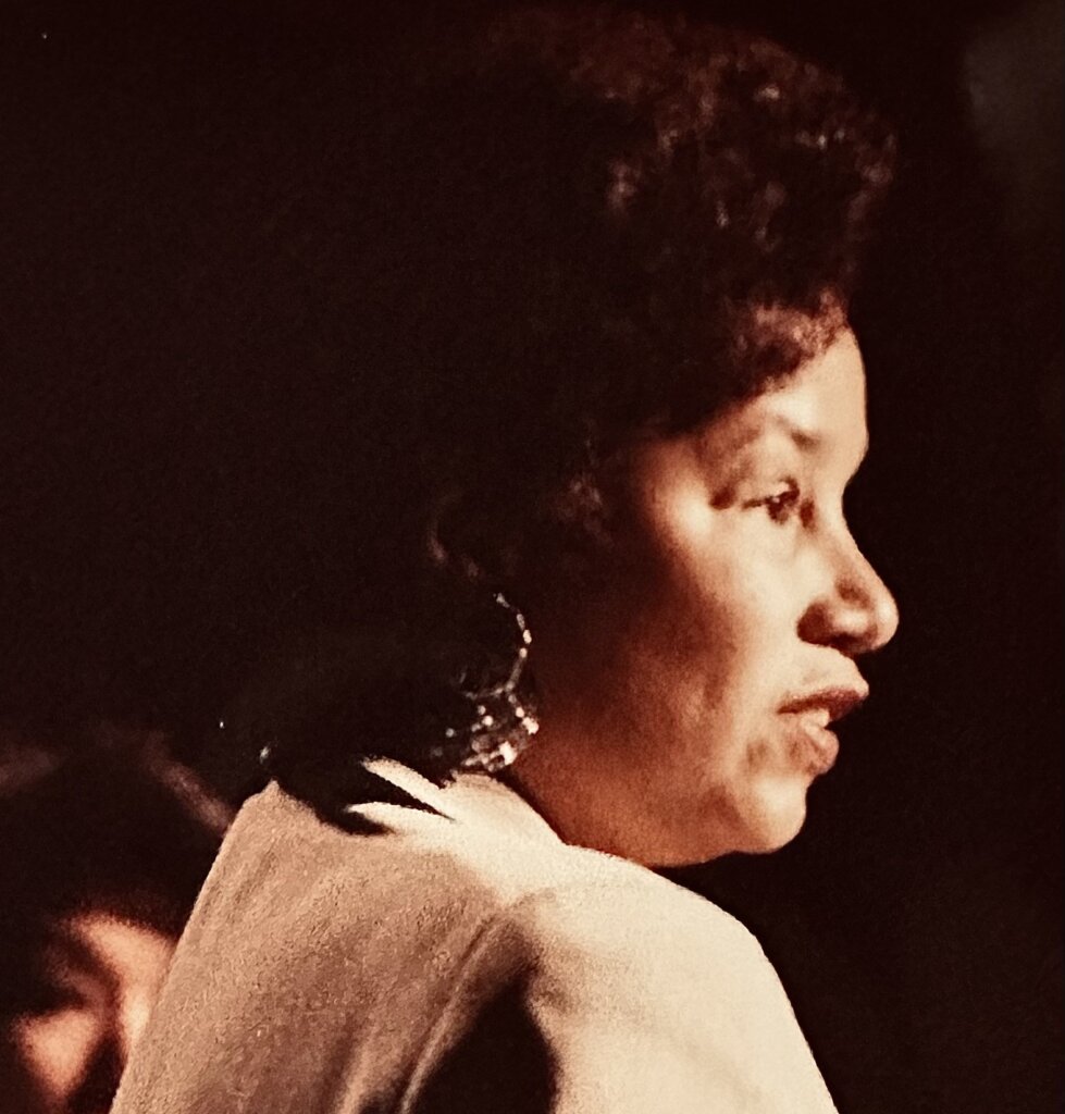 A profile photo of Nancy Hicks Maynard, a Black woman, speaking during the 1980s. She is lit from the front by a stage light or spotlight. She wears large dangling earrings and a suit jacket with shoulder pads and has short, dark curly hair coiffed around her head.
