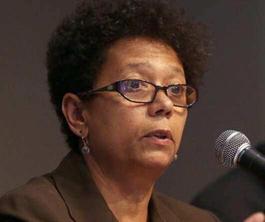 A photo of Dori J. Maynard, a Black woman, wearing glasses and a dark brown suit and speaking into a microphone. She has very short curly hair.