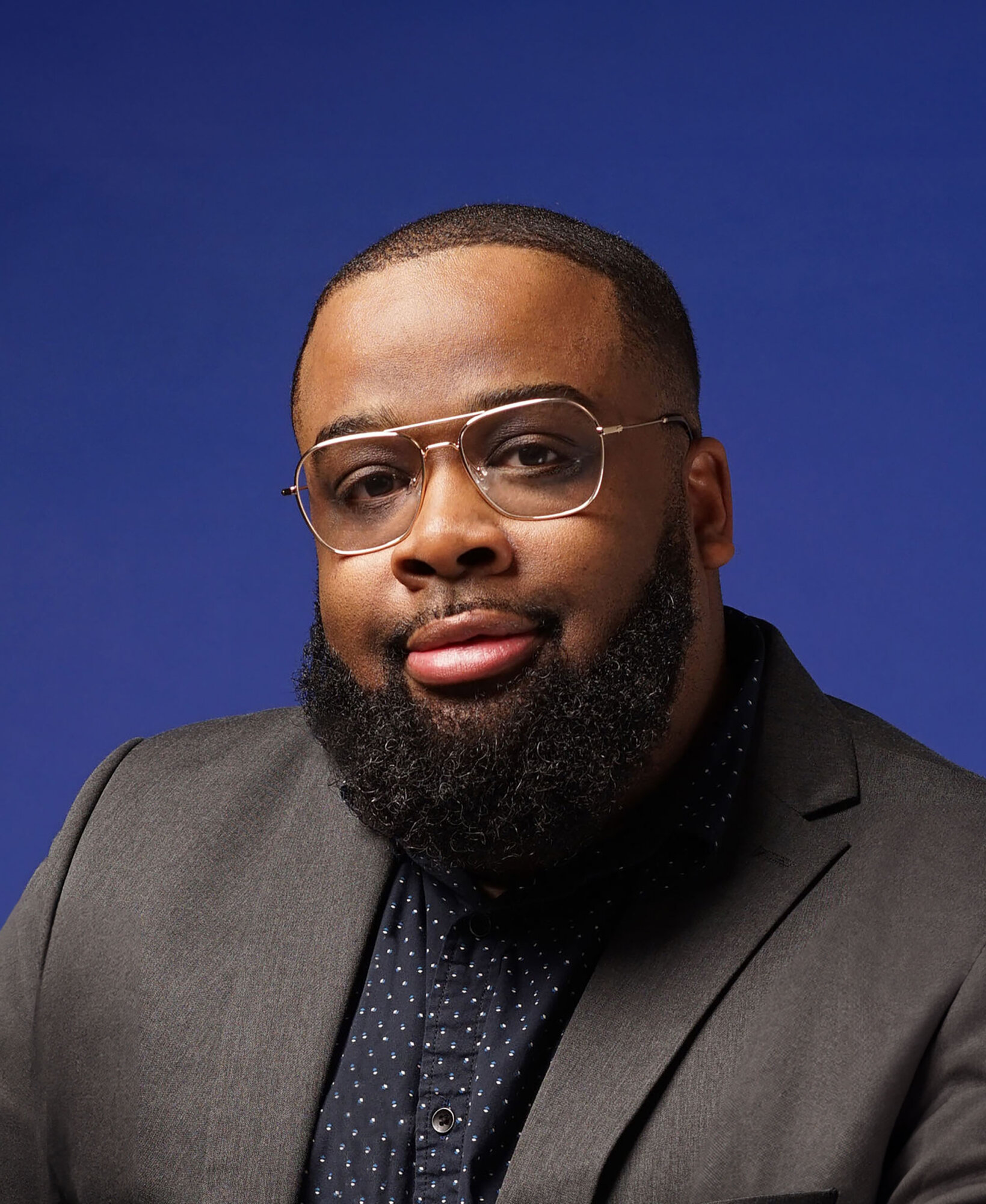 A professional headshot of Mark S. Luckie, a Black man with close cropped hair worn in a short tapered fade, wire-rimmed glasses, and a full, rounded beard. He wears a gray suit jacket and a dark blue button-down shirt.