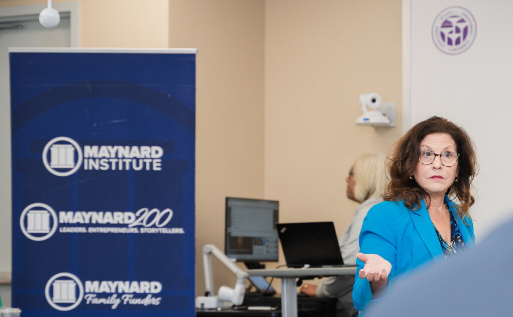 A photo of Marisa Porto, who wears a bright blue blazer and tortoise shell glasses. She has dark brown curly hair that falls to her shoulders and gestures while speaking. Behind her, a dark blue standing banner has white Maynard Institute branding.