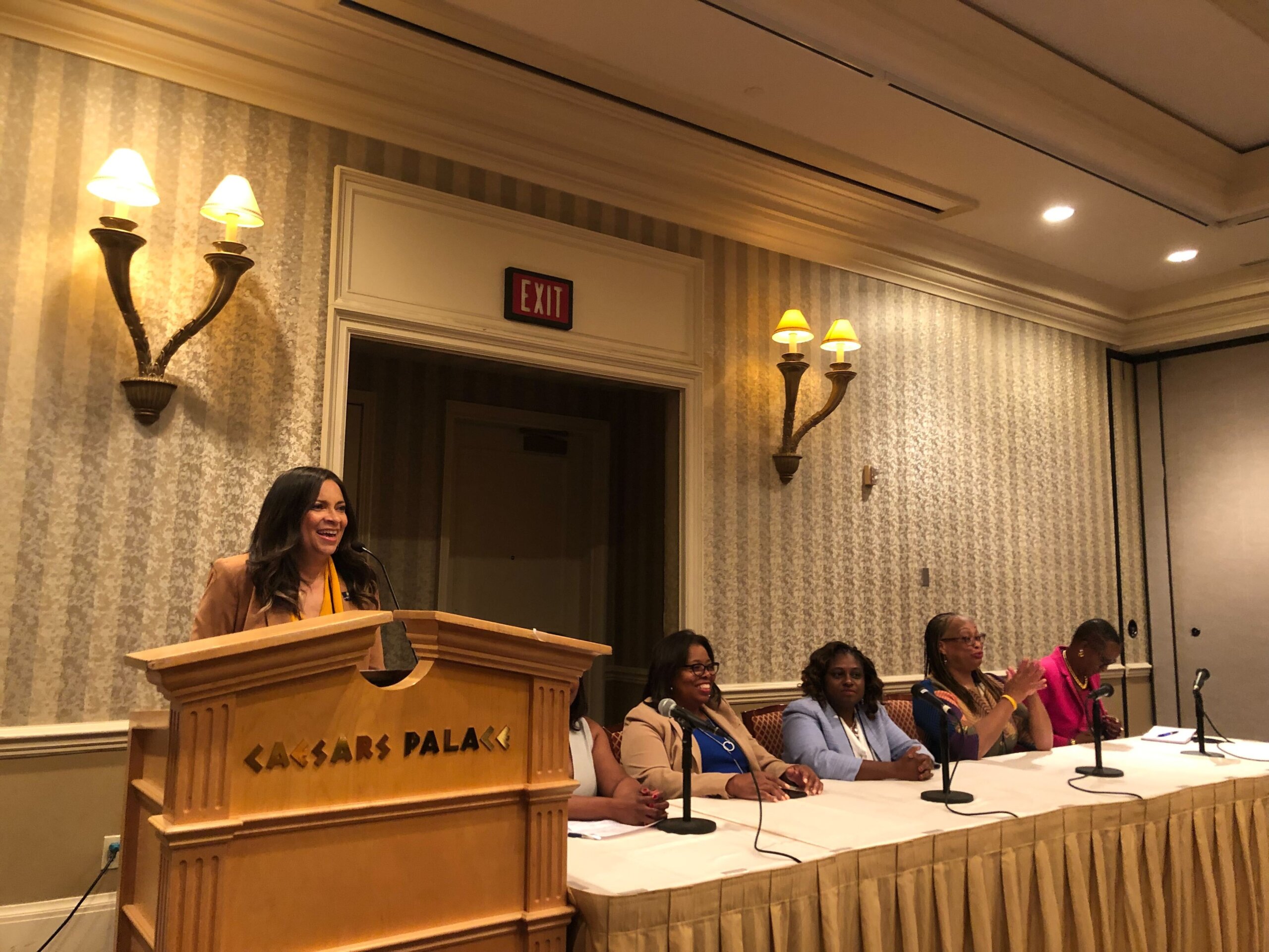 In a large hotel ballroom/conference room, the camera faces a panel of Black women, in colorful yet professional dress. One woman to the far left wearing a camel suit and yellow blouse stands at a podium that reads "Caesar's Palace." Five other women on the panel sit at a long table, small microphones on stands in front of each of them.