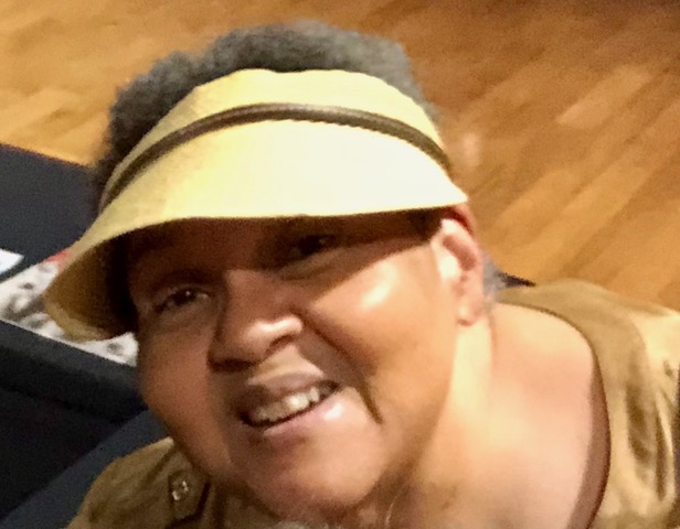 A black woman looks up at the camera. She wears a khaki shirt and a visor and has graying short afro hair.