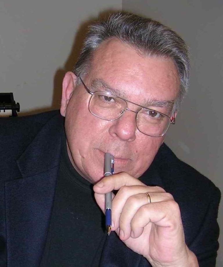 A photo of a white man with graying hair who wears a dark suit and holds a pen to his lips. He wears wire-rimmed glasses and smirks at the camera. He also has a plain gold wedding band on his ring finger.