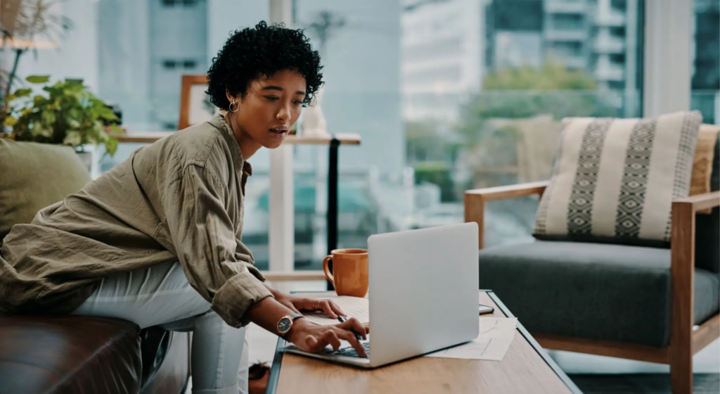 A woman with a medium complexion, Asian features and dark curly hair leans forward to type on a laptop that sits on a coffee table before her. She sits on a dark leather couch in what looks like a small seating area in an office. She wears a sage green linen blouse and hoop earrings, and behind her an orange mug sits on the table.