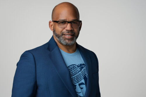 A photo of Martin G. Reynolds, Co-Executive Director of the Maynard Institute. A Black man, he wears glasses, is bald, has a graying beard, and wears a sharp blue suit jacket over a light blue t-shirt with a dark blue graphic. Behind him is a light gray background.