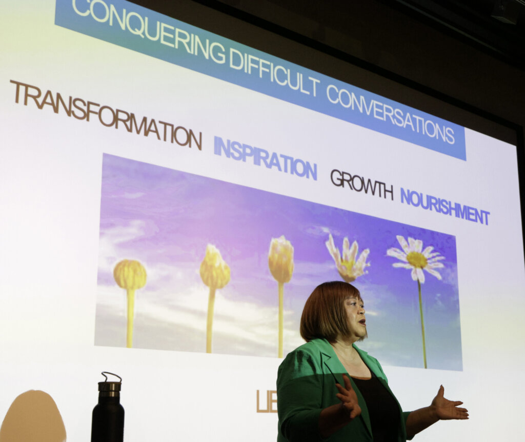A Black woman with short light brown hair and a green blazer gestures while speaking. Behind her, a large projection screen shows a photo of a small daisy in several stages of blooming. In a light blue banner at the top of the projection white text reads 