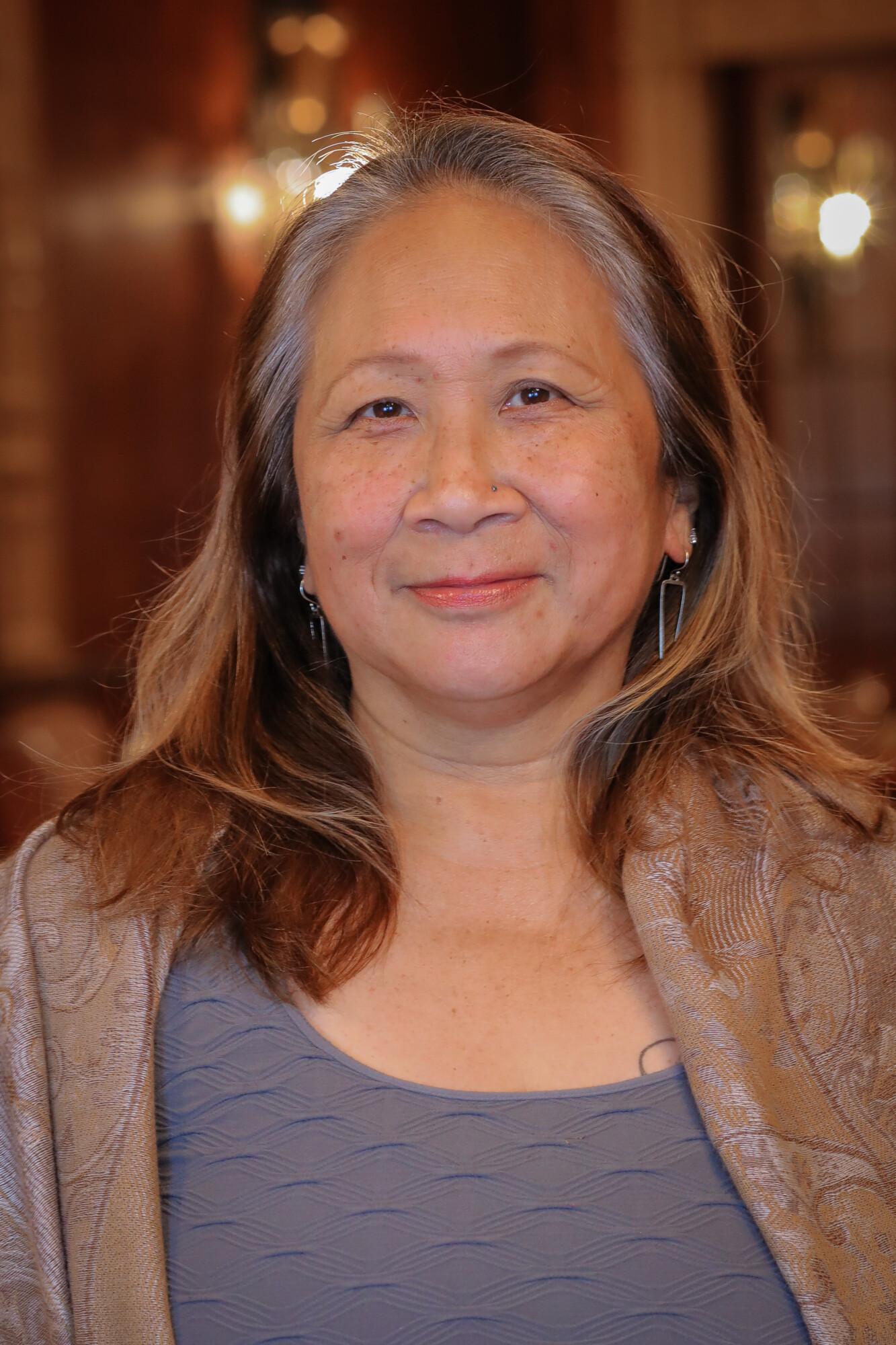 A photo of Alida Birnam, an Asian woman with brown hair graying at the temples. She smiles and wears silver earrings and a pashmina.