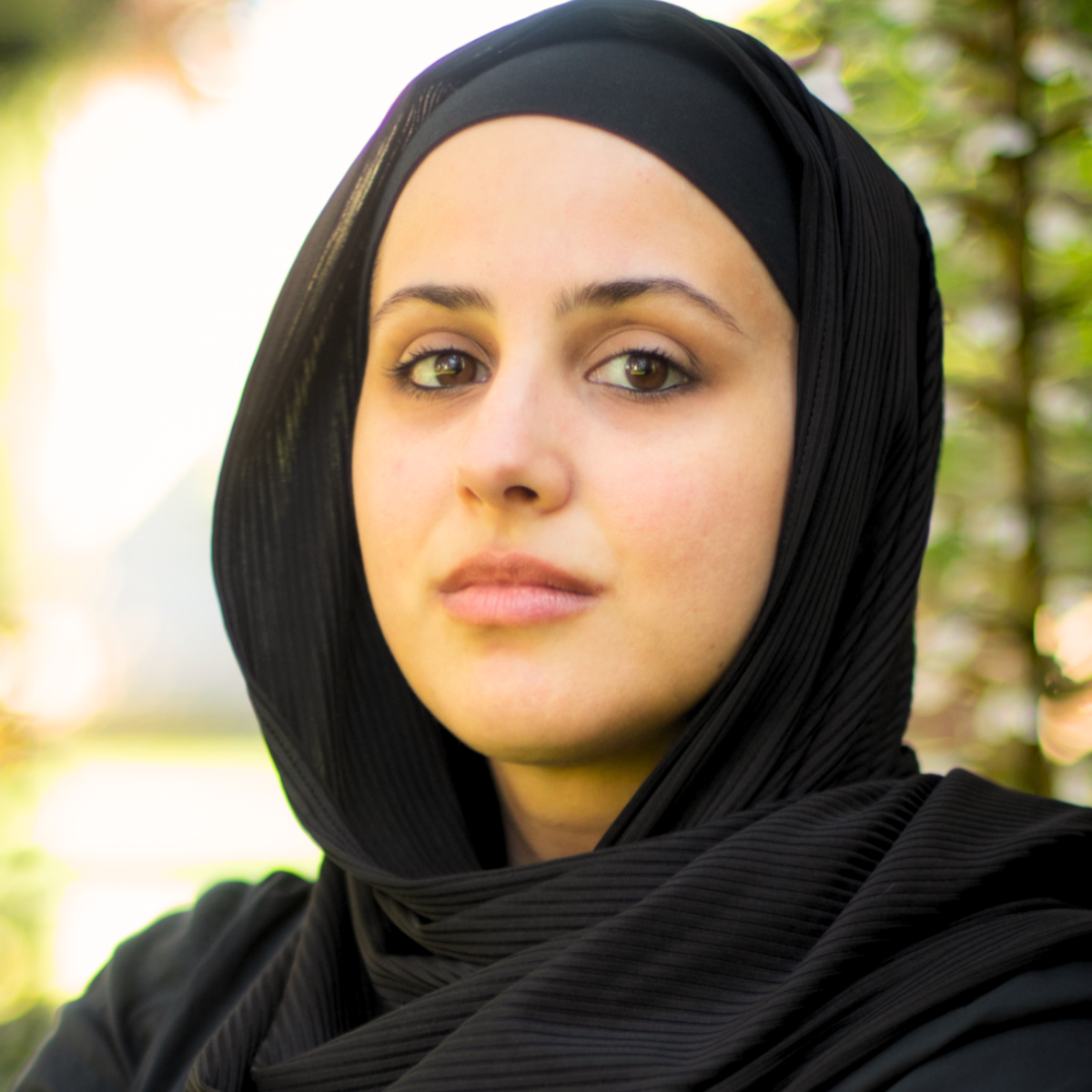 A photo of Amani Hamed, she wears a black hijab and black shirt. Behind her are blurry background trees.
