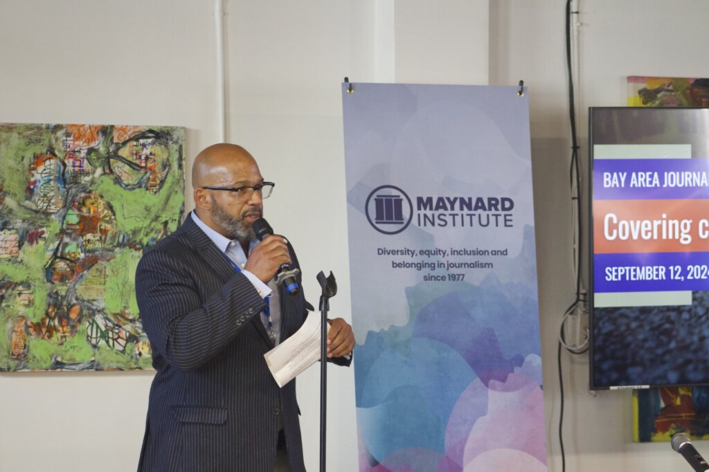 Martin Reynolds, a Black man, is bald and wears glasses and a pinstriped blue suit with a light blue shirt. He has a graying beard and holds a microphone and piece of paper. Behind him, abstract artworks adorn the walls and a standing banner with a collage background has the Maynard Institute logo and dark blue text that reads 