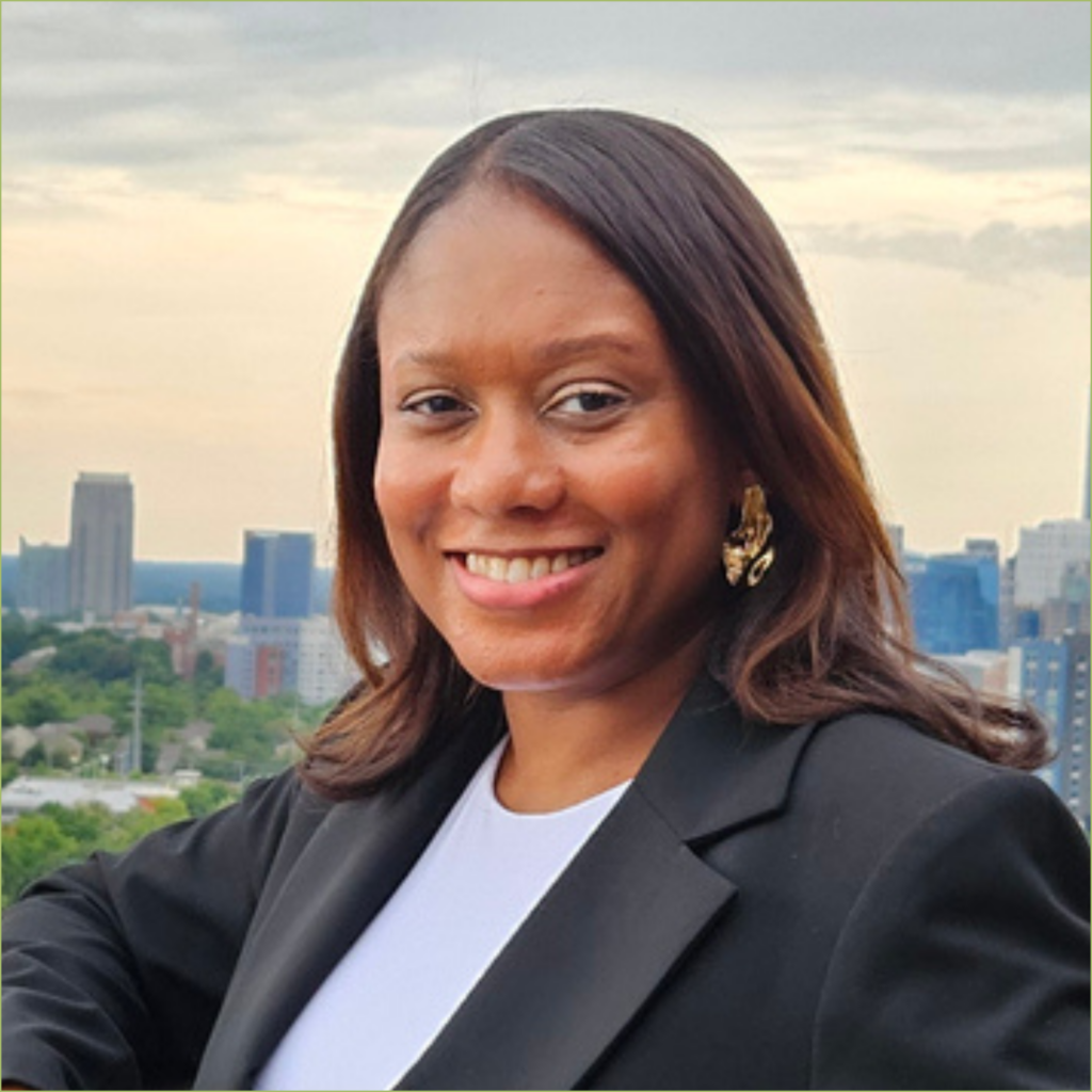 A photo of Nadia Campbell-Mitchell, a Black woman with shoulder-length hair wearing a black blazer and light blue shirt with gold earrings. She leads against a railing with one arm up, behind her is a city skyline.