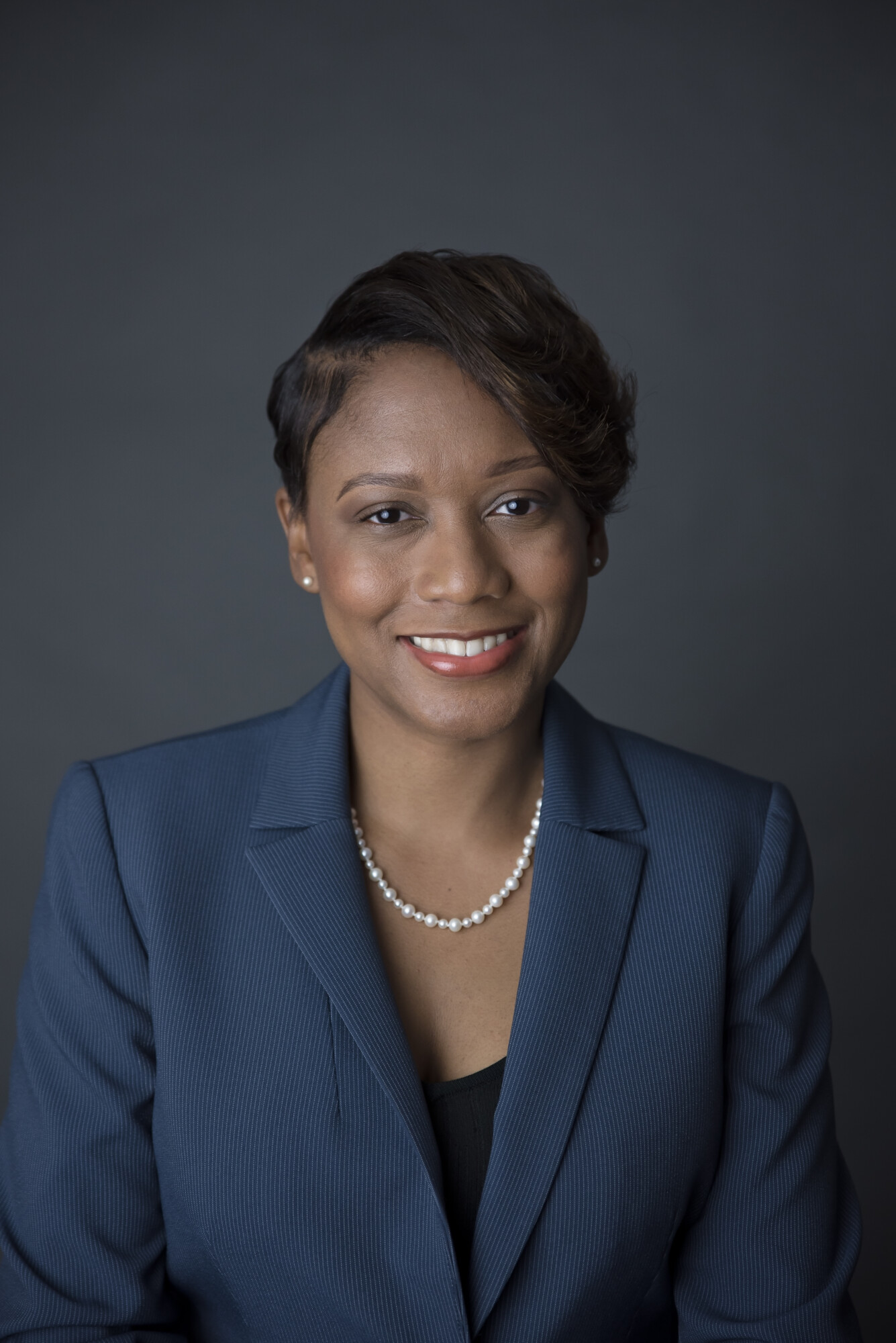 A professional photo of Nadia Campbell-Mitchell, a Black woman, who wears a dark blue suit jacket, black shirt, and pearl necklace and earrings. Behind her is a dark gray background. Her hair is short and resembles a pixie cut.