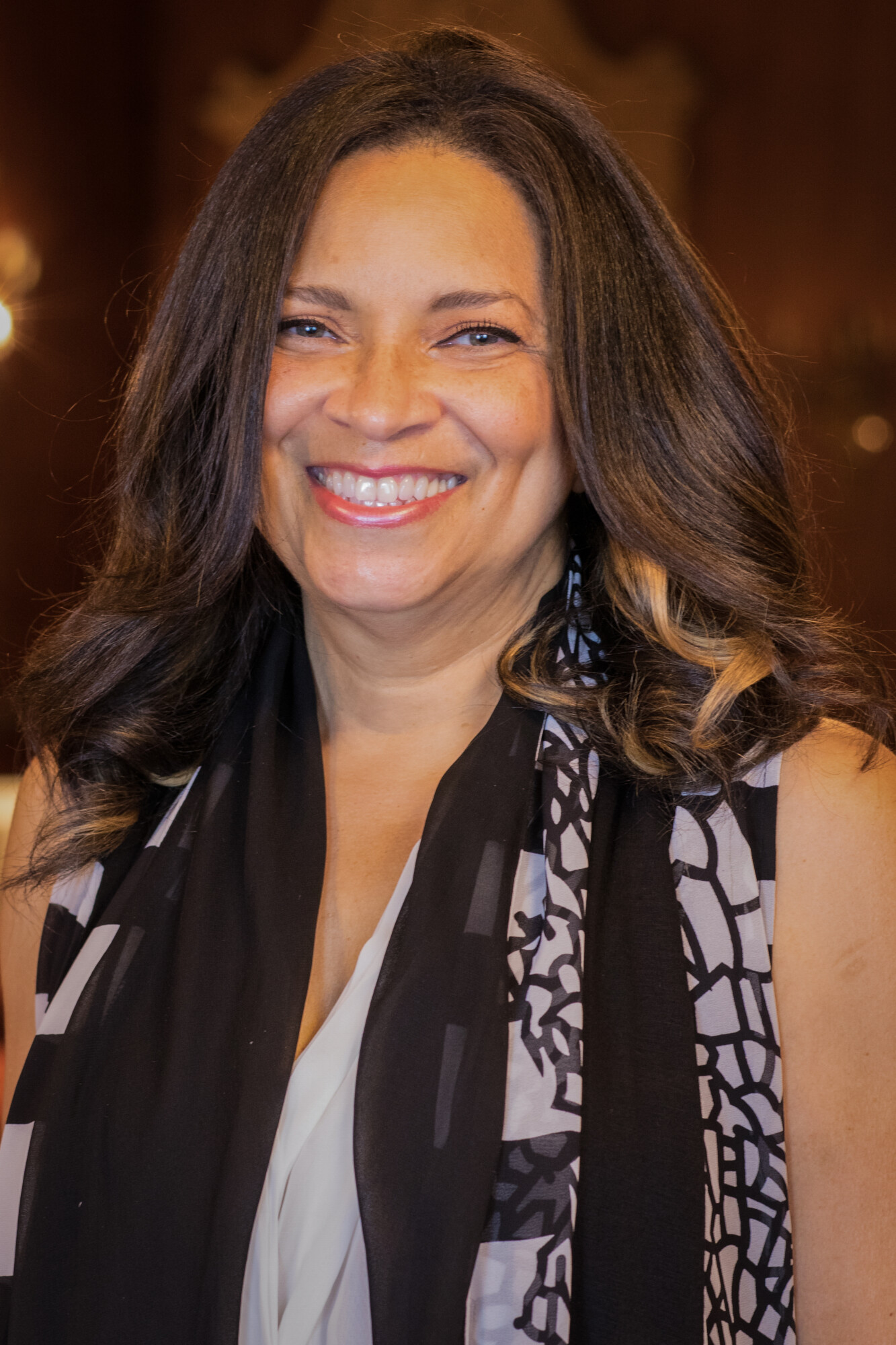 A photo of Felecia D. Henderson, a Black woman with a honey complexion and shoulder-length brown hair with loose curls and highlights. She smiles and wears a white blouse and black and white scarf.