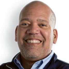 A photograph of Christian Hendricks, a Black man with light skin, blue eyes and a shaved head, smiling and wearing a blue button down shirt and dark blue jacket.