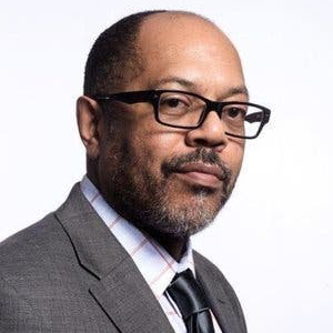 A photo of Kevin Merida, a Black man with black glasses, a short beard, and a gray suit with a white shirt striped with red and a black tie.