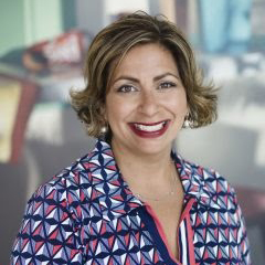 A photo of Kim Bardakian, a woman with a colorful geometric shirt, a short hairstyle and blonde highlights, smiling to the camera.