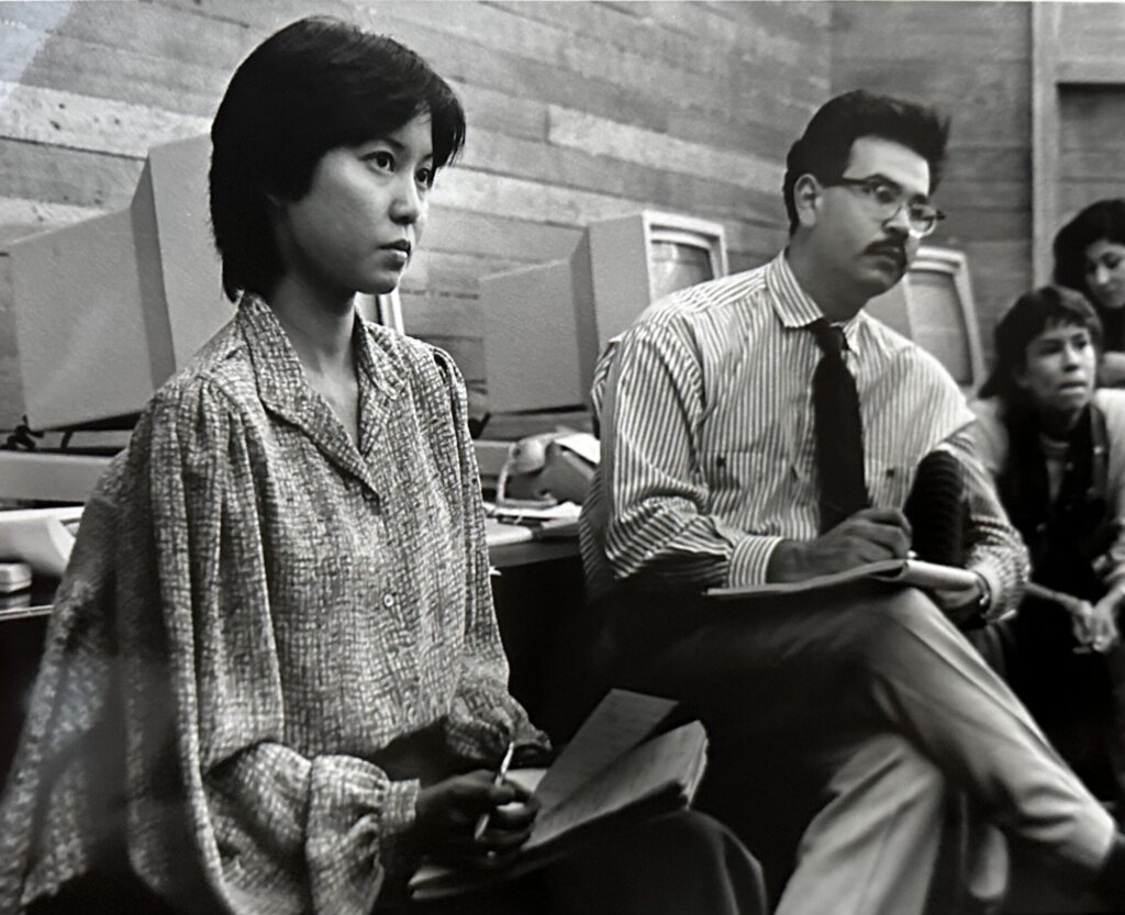 A black and white photo. A young Asian woman sits closest to the camera wearing a loose collared blouse and writing on a notepad while listening intently to someone out of frame. Behind her, a man with dark hair, glasses, and a mustache also takes notes. He wears a striped shirt and a tie. Behind him several more people listen to the off-screen person. They sit in front of desks with a row of boxy computer monitors. From the computers, clothing, and hairstyles this photo appears to have been taken in the mid-1980s.