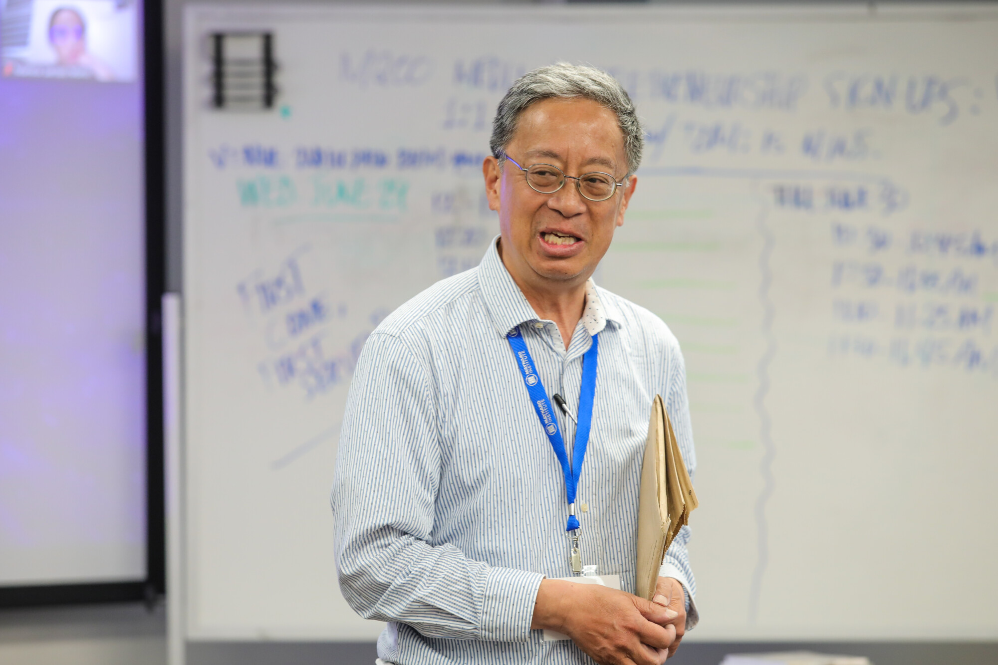 A photo of Dickson Louie, an Asian man with graying hair, glasses, and a blue lanyard with the Maynard Institute logo. He holds several manila folders. Behind him a whiteboard is covered with what appear to be numbers and figures in the blurred background.