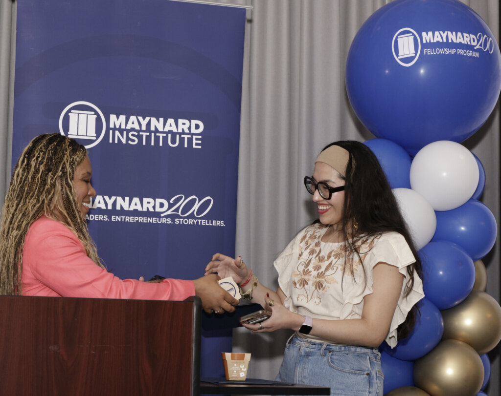 Left, a young black woman with dark braids and blonde highlights wears a bright pink blazer and stands before a podium, she hands something to a young woman with thick black glasses, a tan headband, and waist-length dark wavy hair. A banner and balloon tower behind them are branded with the Maynard Institute logo in white and the Maynard Institute's signature dark blue color.