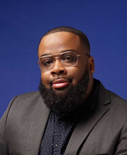 A professional headshot of Mark S. Luckie, a Black man with close cropped hair worn in a short tapered fade, wire-rimmed glasses, and a full, rounded beard. He wears a gray suit jacket and a dark blue button-down shirt.