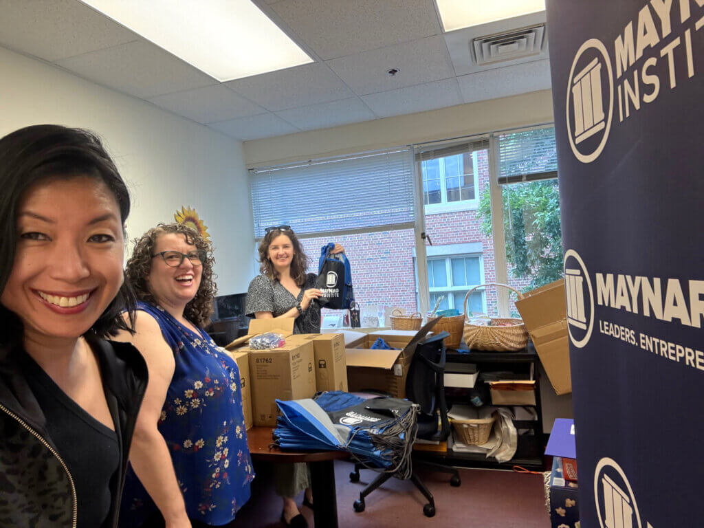 In the foreground, Odette Alcazaren-Keeley, an Asian woman with dark shoulder-length hair wearing a black shirt and jacket, takes a selfie. Before her is a dark blue standing banner with the Maynard Institute logo in white. Behind her two white women smile , one wants in front of and the other behind a table laden with Maynard Institute-branded tote backs. The woman behind the table holds one up. They appear to be in an office supply room.