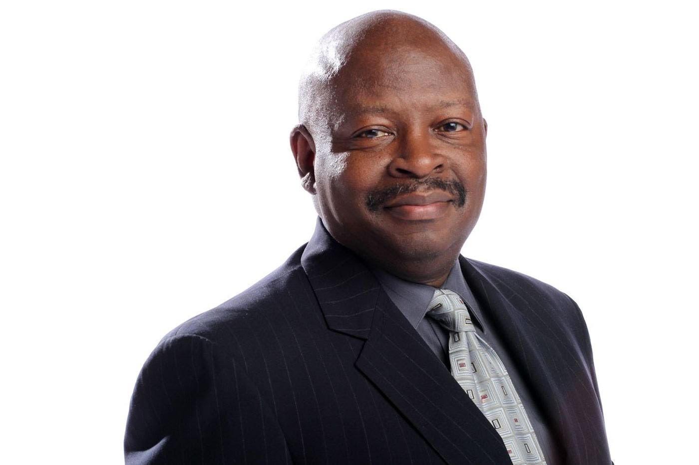 A photo of Michael Days, a bald Black man with a mustache wearing a black suit jacket, blue-gray shirt and silver/geometric tie. He smiles subtly.