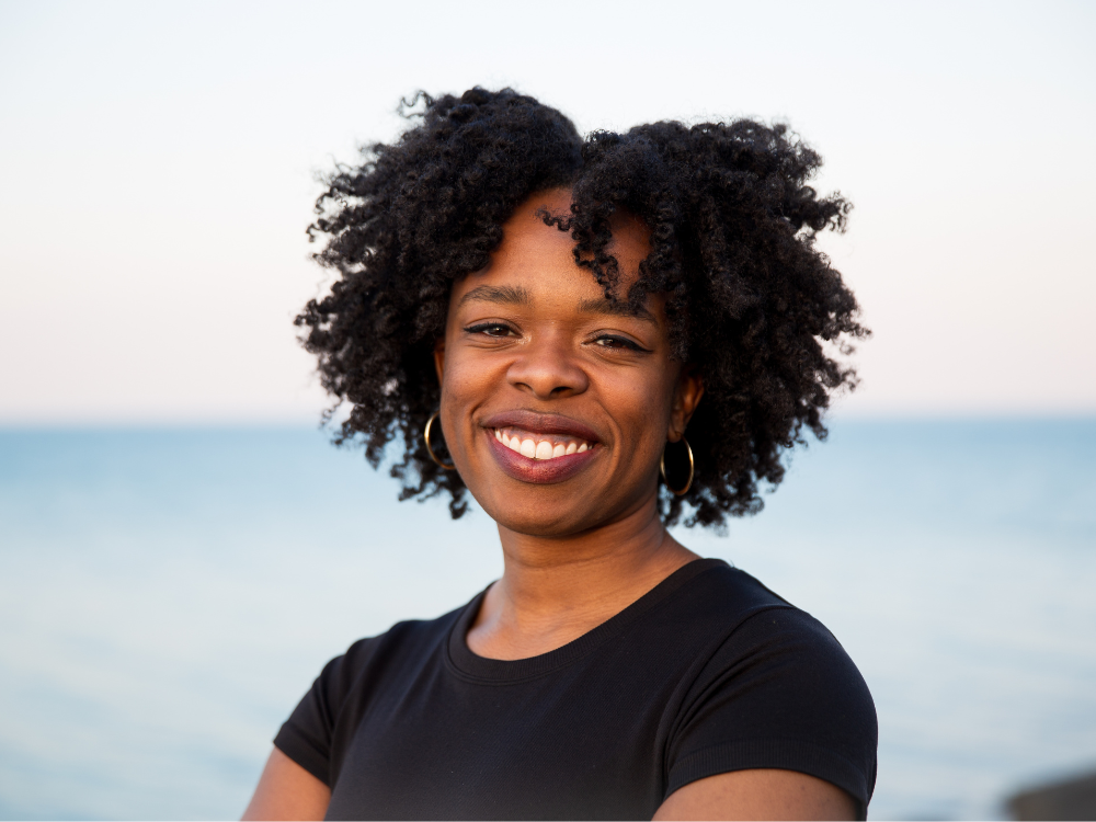 A photo of Jasmine Barnes, a Black woman with tight curly hair parted n the middle. She wears a black short sleeved shirt and smiles broadly. Behind her is the ocean.
