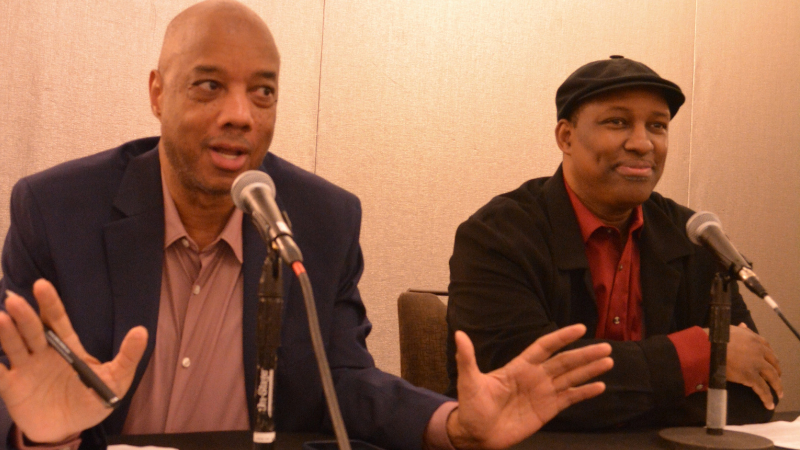 A photo of two Black men seated side by side at a table. Each has a microphone on a small stand in front of him. At left: CAPTION: Gromer Jeffers Jr., political reporter at The Dallas Morning News, a Black man who is bald and wears a blazer and button-down shirt, and Will Ford. a Black man with a flat cap and red button down shirt with dark jacket.