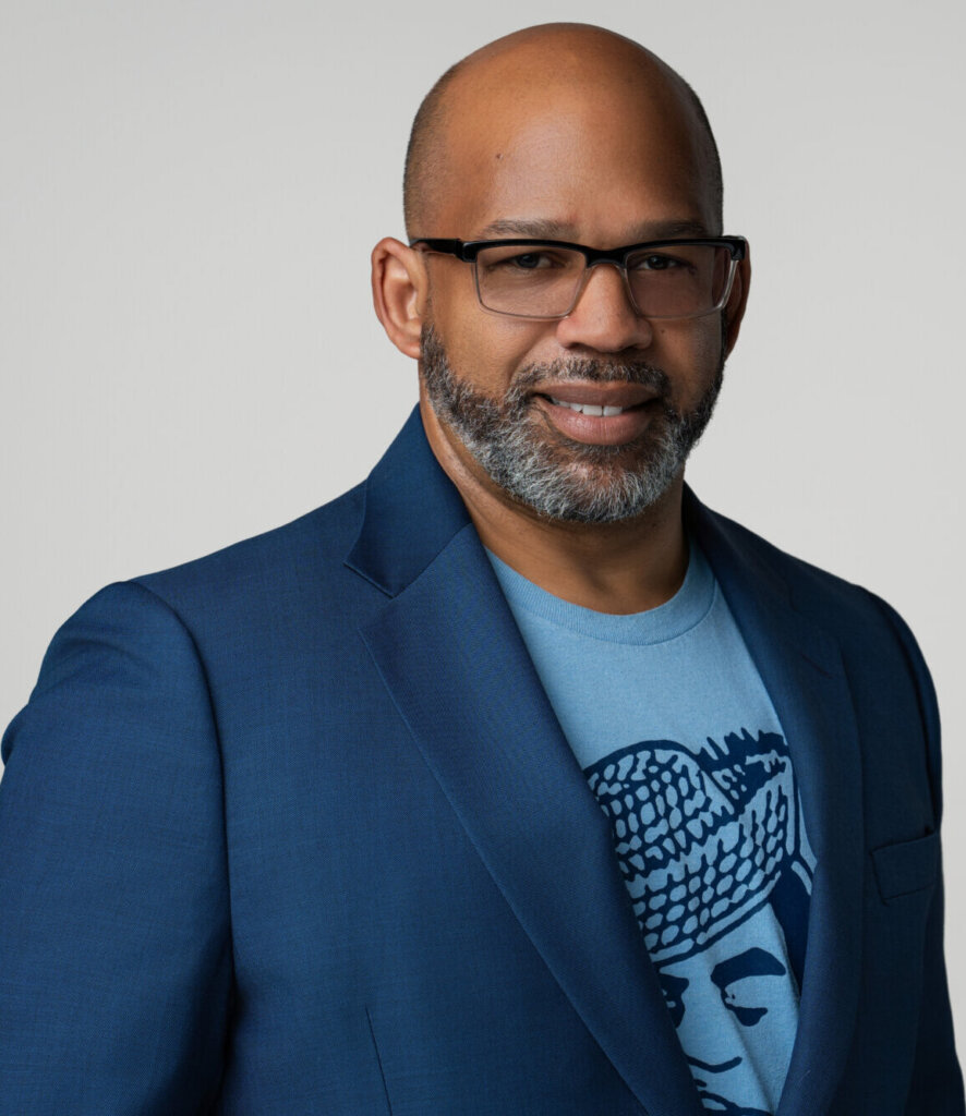 A photo of Martin G. Reynolds, Co-Executive Director of the Maynard Institute. A Black man, he wears glasses, is bald, has a graying beard, and wears a sharp blue suit jacket over a light blue t-shirt with a dark blue graphic. Behind him is a light gray background.